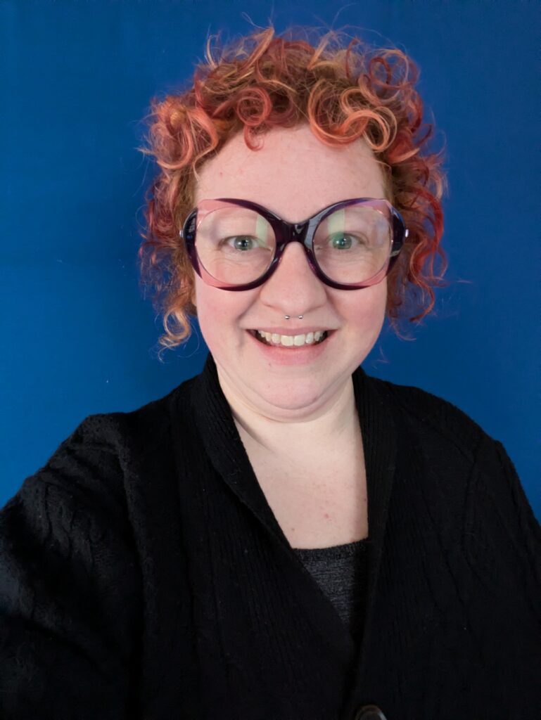 Image of Heather O'Shea in front of a blue backdrop. Heather has short, red, curly hair, pale skin, and green eyes. She is wearing a black top and large, purple, round-framed glasses. 
Click the image to read Heather's story of how the Green Bean Collective came to be. 